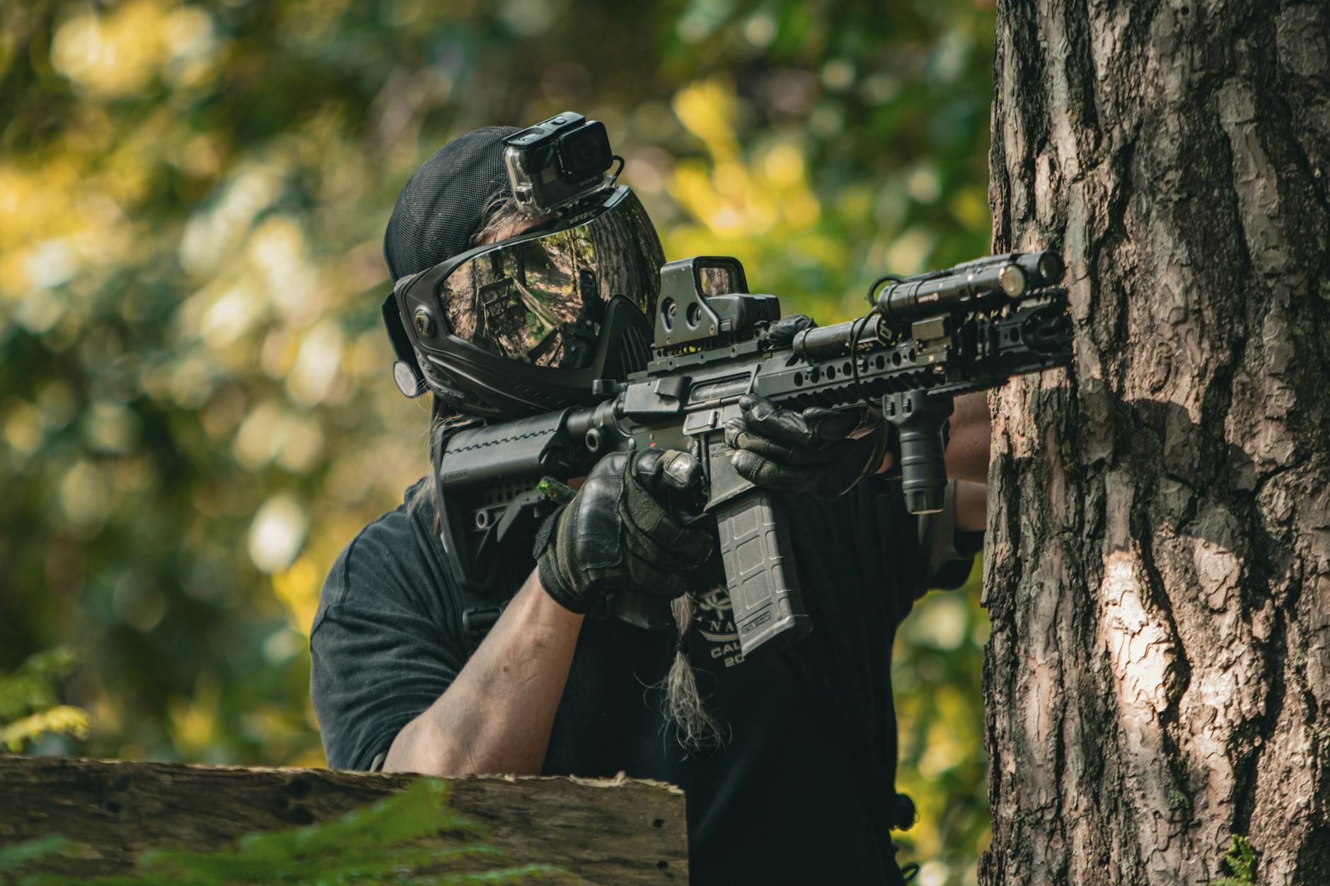 man in protective paintball mask aiming a weapon hiding behind a tree